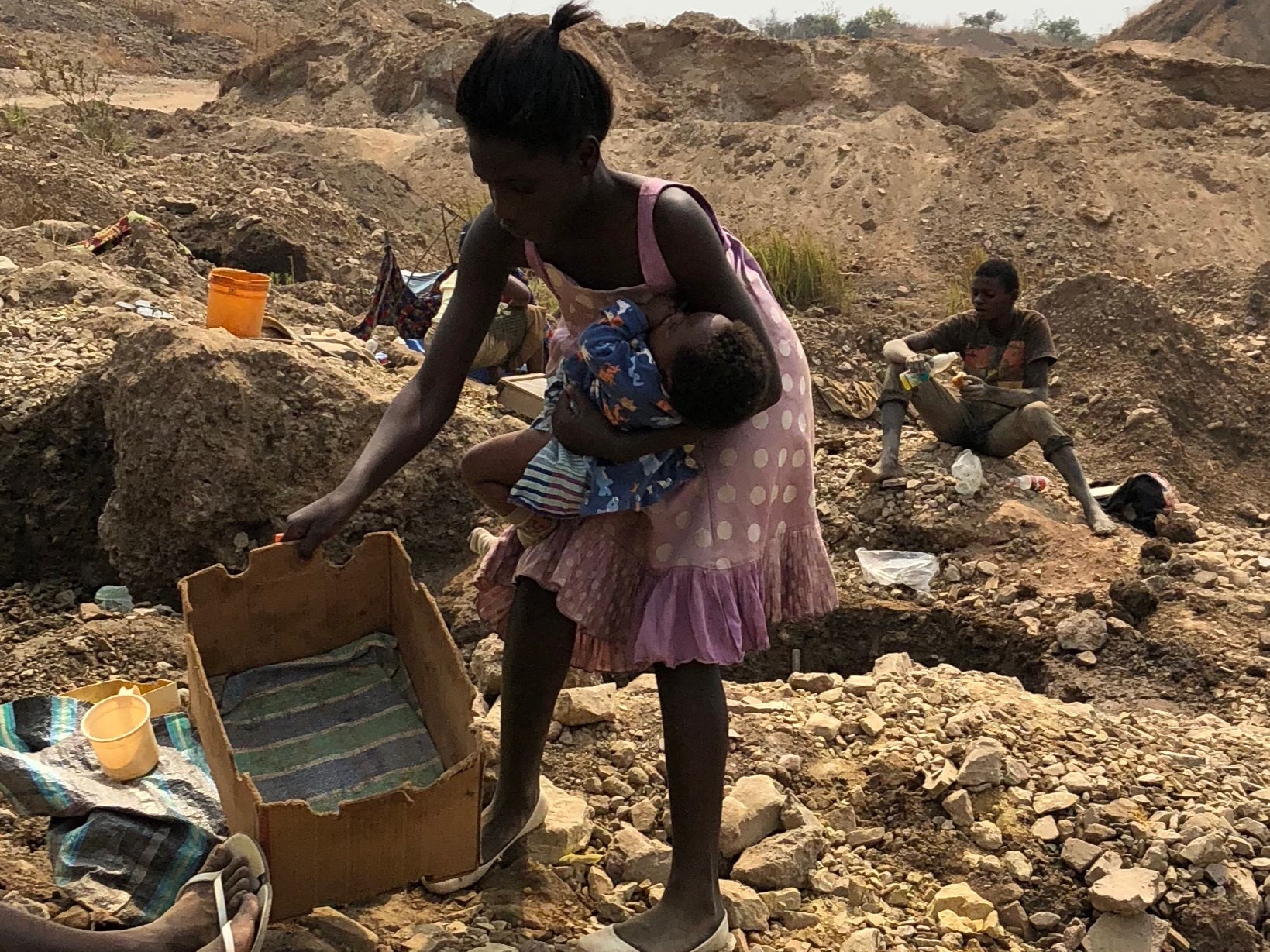 <p>A woman places a baby into a cardboard crib on the ground while she works at a cobalt mine site. Women are paid only a fraction of the paltry sums paid to men for their mining work</p>