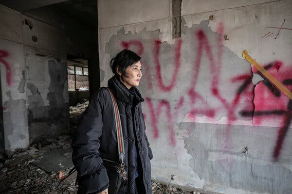 A woman with a sad expression stands in a dilapidated building with graffiti and debris in the background.
