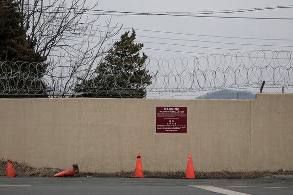 A wall topped with barbed wire and a sign to keep out. In the foreground are asphalt and orange traffic cones.