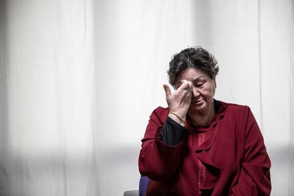 A woman sitting against a blurry white background wipes her eyes.